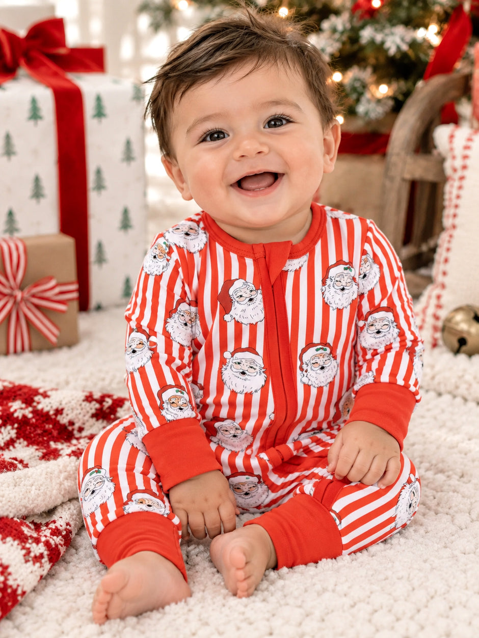 Toddler in festive striped pajamas with Santa faces, smiling on a soft rug surrounded by holiday gifts.
