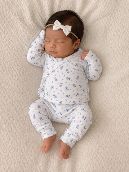Sleeping newborn dressed in a floral onesie, wearing a white bow headband on a soft, textured blanket.