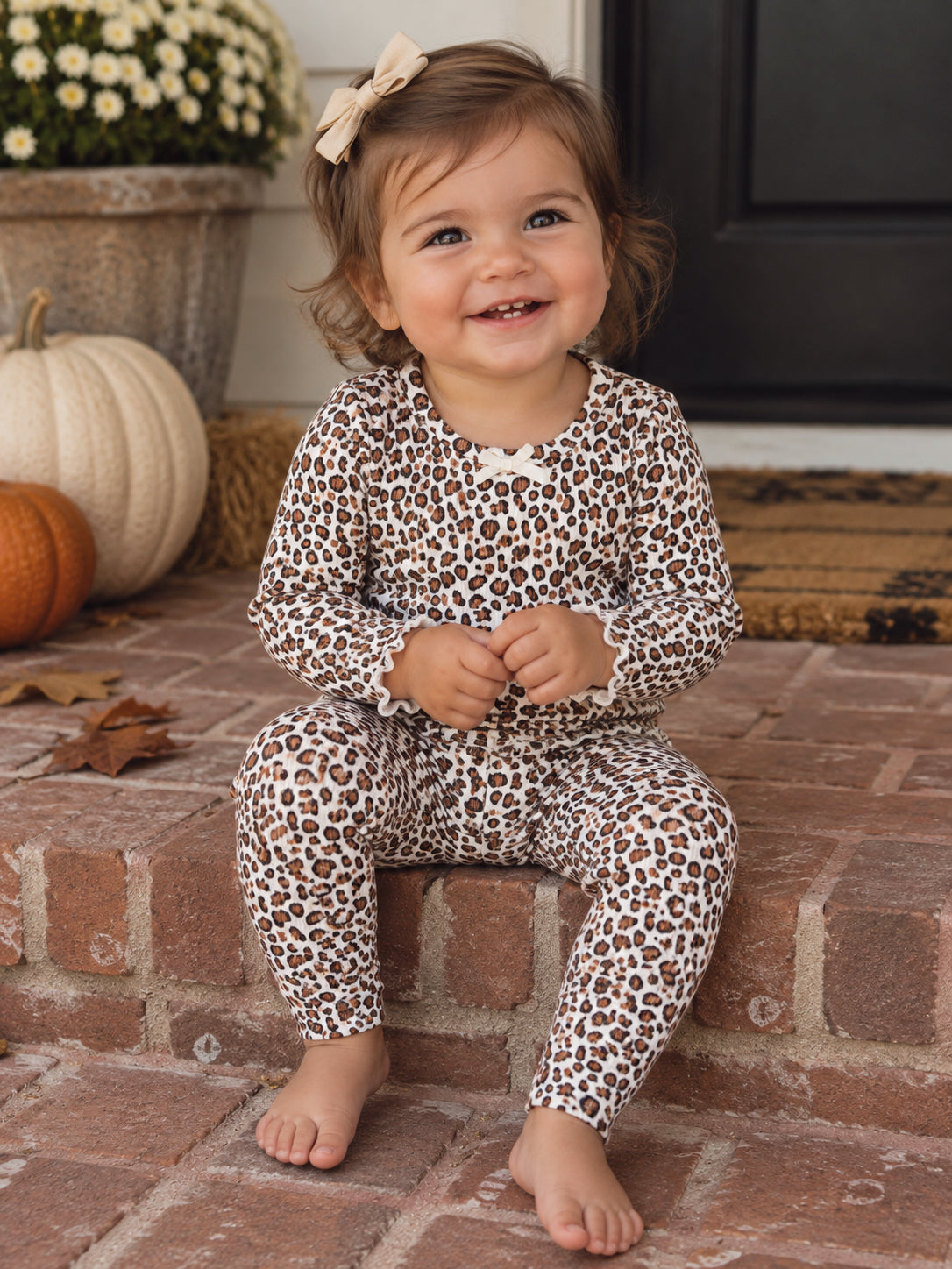 Smiling toddler in leopard print outfit sits on brick steps, surrounded by pumpkins and flowers for fall decor.
