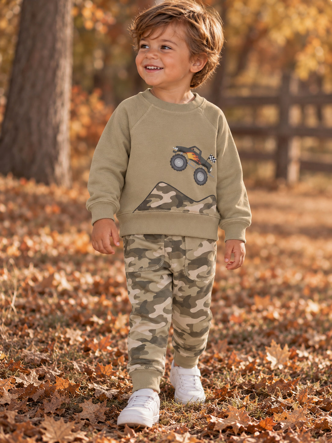 Smiling boy in a gray sweater and camo pants stands on leaves, with a toy truck graphic on his shirt.