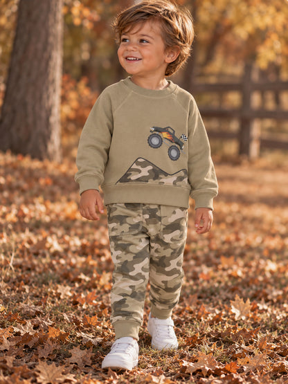 Smiling boy in a gray sweater and camo pants stands on leaves, with a toy truck graphic on his shirt.
