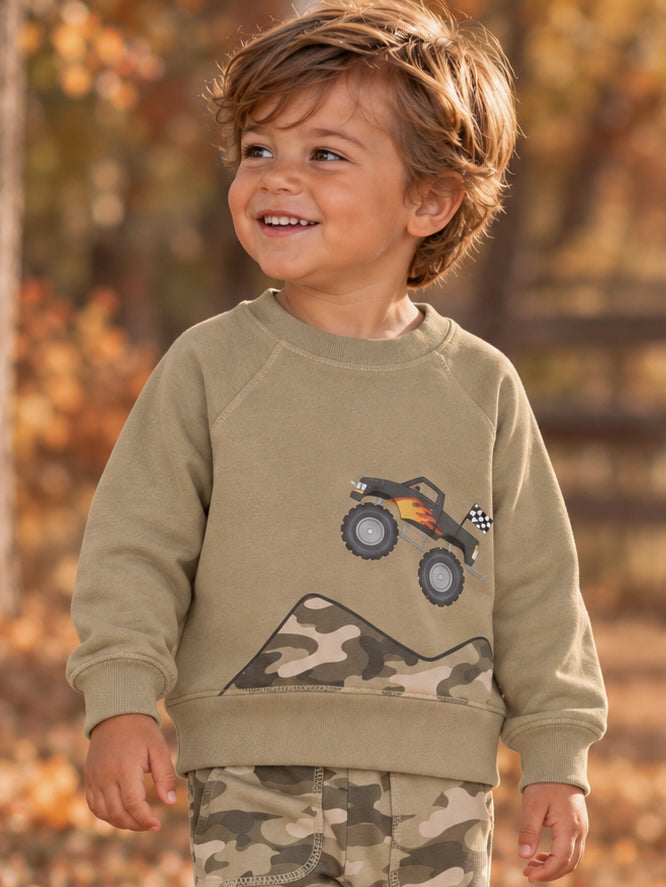 Smiling boy in a khaki sweatshirt with a monster truck graphic, surrounded by autumn foliage.