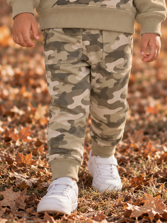 Child in camouflage pants and white sneakers walking through fallen leaves.