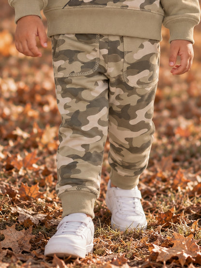 Child in camouflage pants and white sneakers walking through fallen leaves.