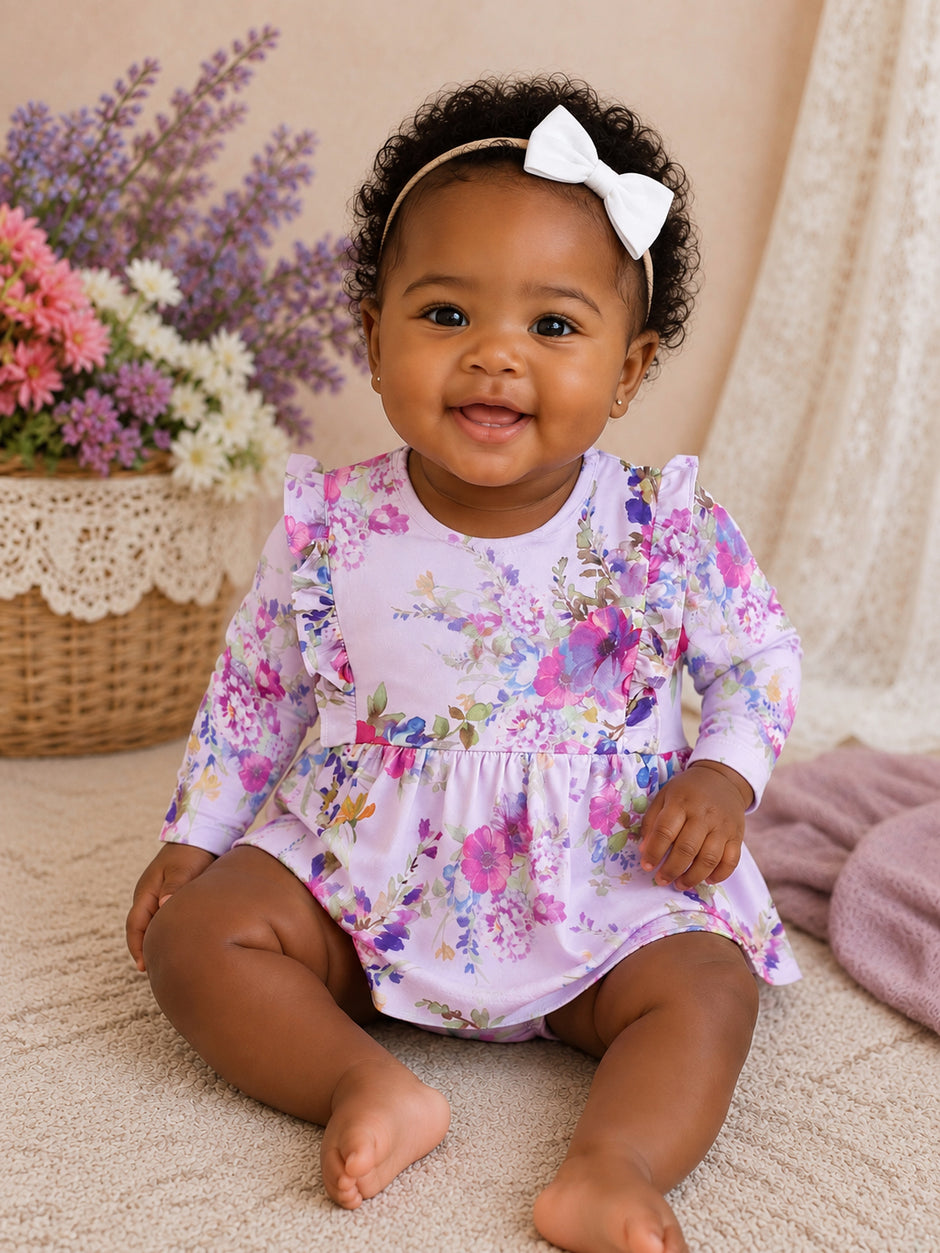 Smiling baby girl in a floral dress and white bow, sitting on a soft surface with flowers in a basket nearby.