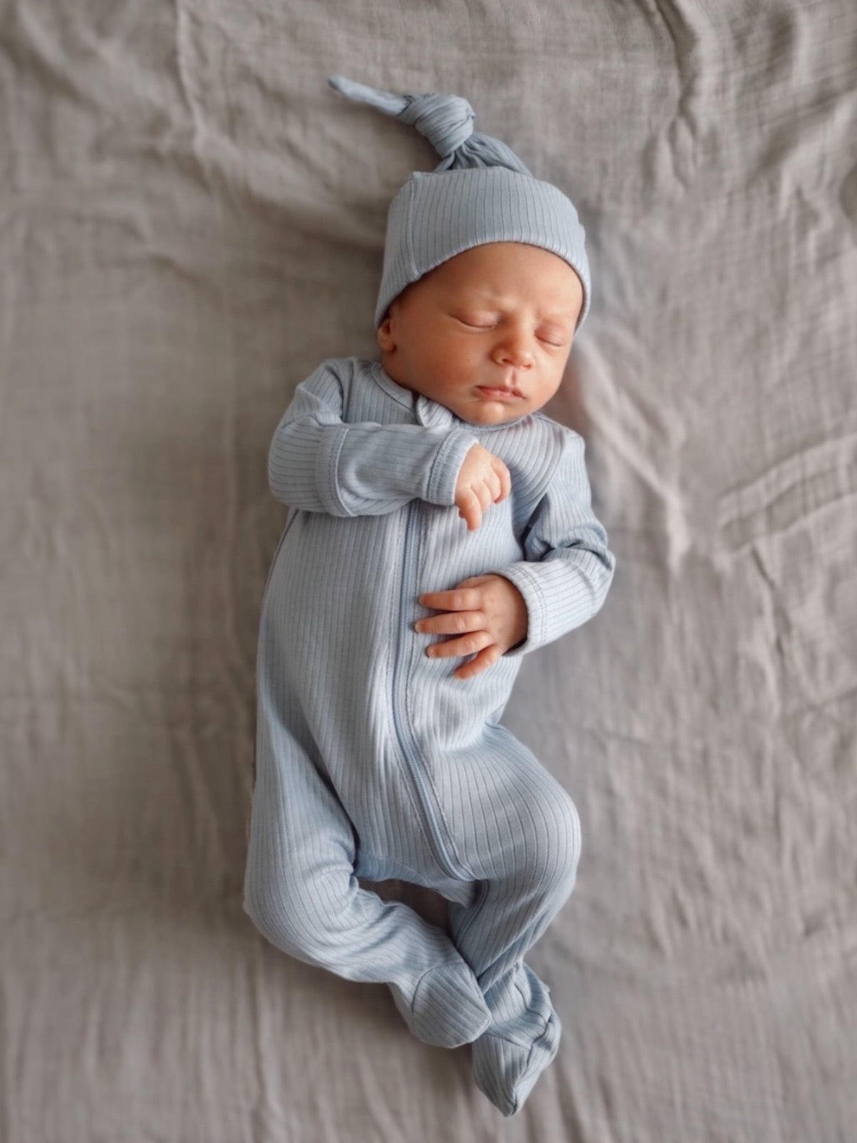 Newborn baby in a light blue outfit and hat, peacefully sleeping on a soft, neutral-colored blanket.