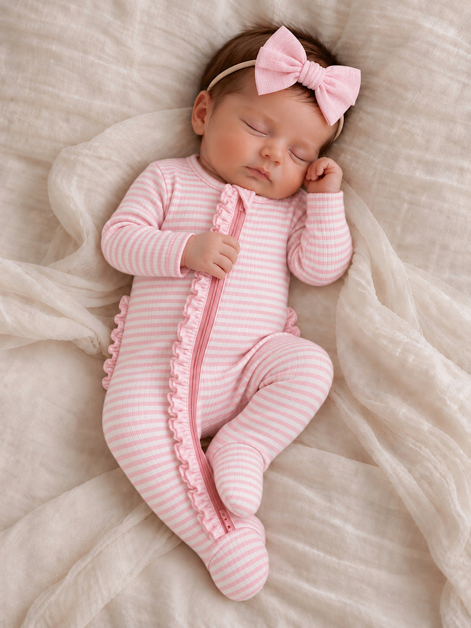 Sleeping newborn wearing a pink striped onesie with ruffles and a matching bow, resting on a soft blanket.
