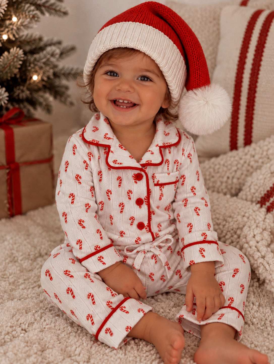 Smiling child in candy cane pajamas and Santa hat, sitting by a decorated Christmas tree and cozy blankets.