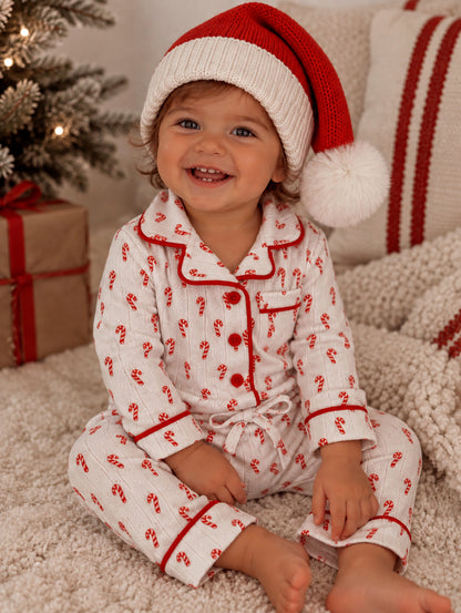 Smiling child in candy cane pajamas and Santa hat, sitting by a decorated Christmas tree and cozy blankets.