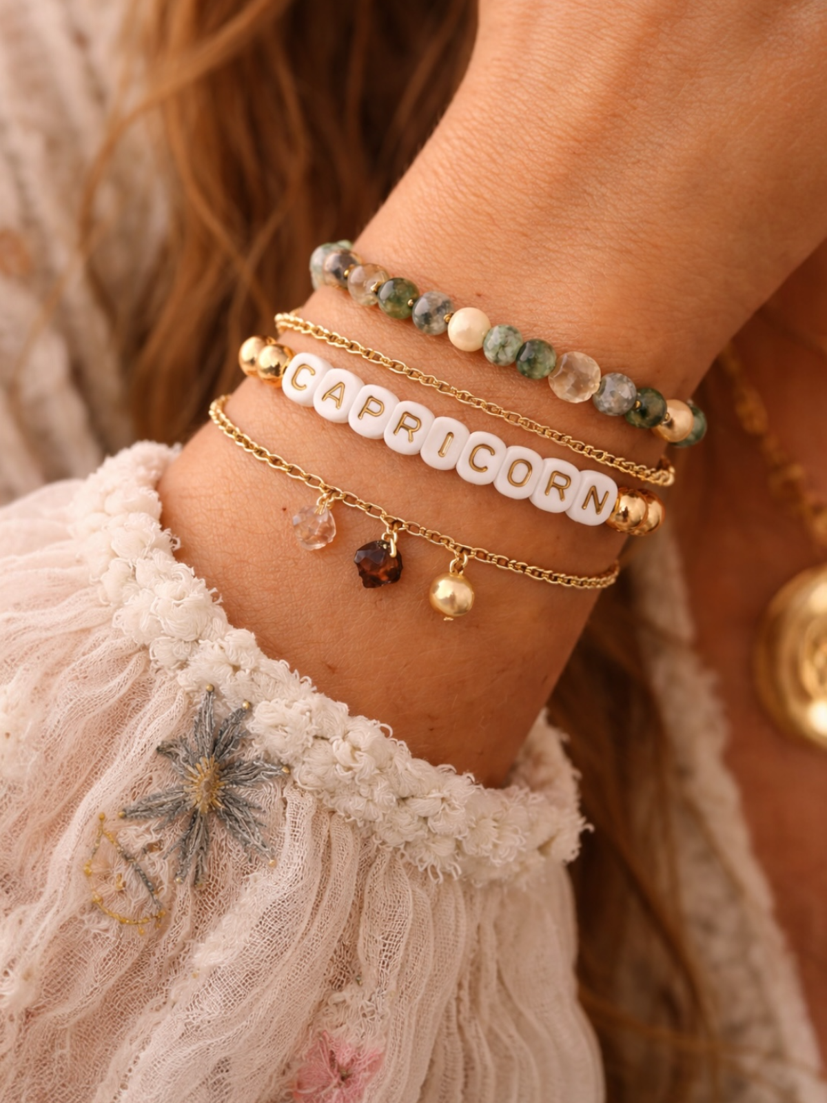 Close-up of a wrist adorned with bracelets, featuring the word "CAPRICORN" and various gemstones on delicate chains.