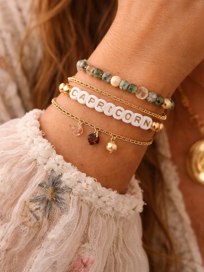 Close-up of a wrist adorned with bracelets, featuring the word "CAPRICORN" and various gemstones on delicate chains.