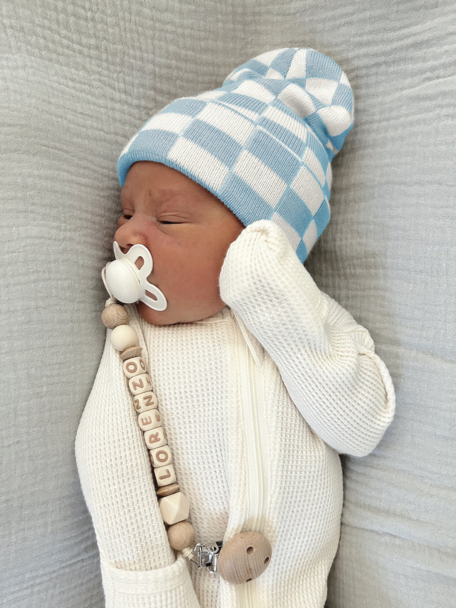 Newborn baby sleeping in a blue and white checkered hat, with a pacifier and a wooden name teether.