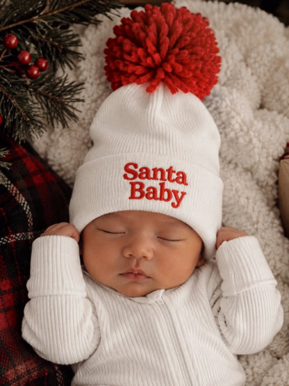 Baby wearing a white "Santa Baby" hat with a red pom-pom, sleeping on a cozy blanket surrounded by festive decor.