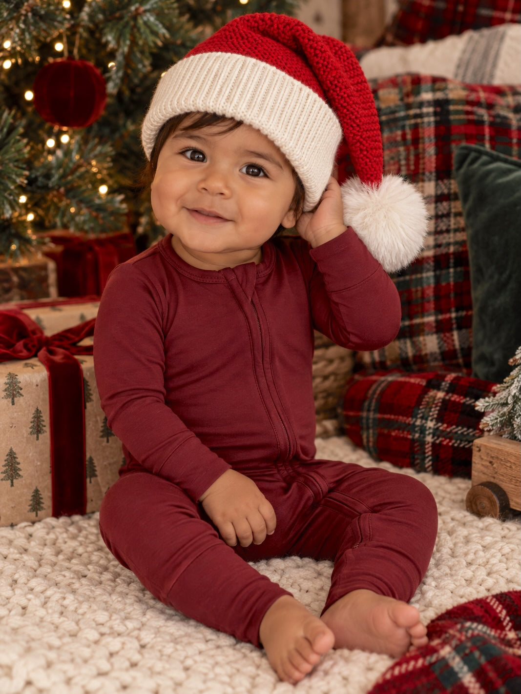 Smiling baby in a red onesie and Santa hat sitting on a cozy, festive blanket with Christmas decorations.