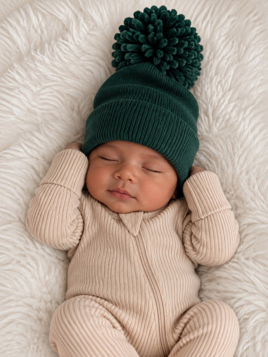 Baby sleeping peacefully on a soft blanket, wearing a cozy green hat with a pompom and a beige ribbed onesie.