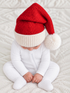 Baby in a knitted red and white Santa hat sits on a cozy blanket, wearing a white outfit, looking down.