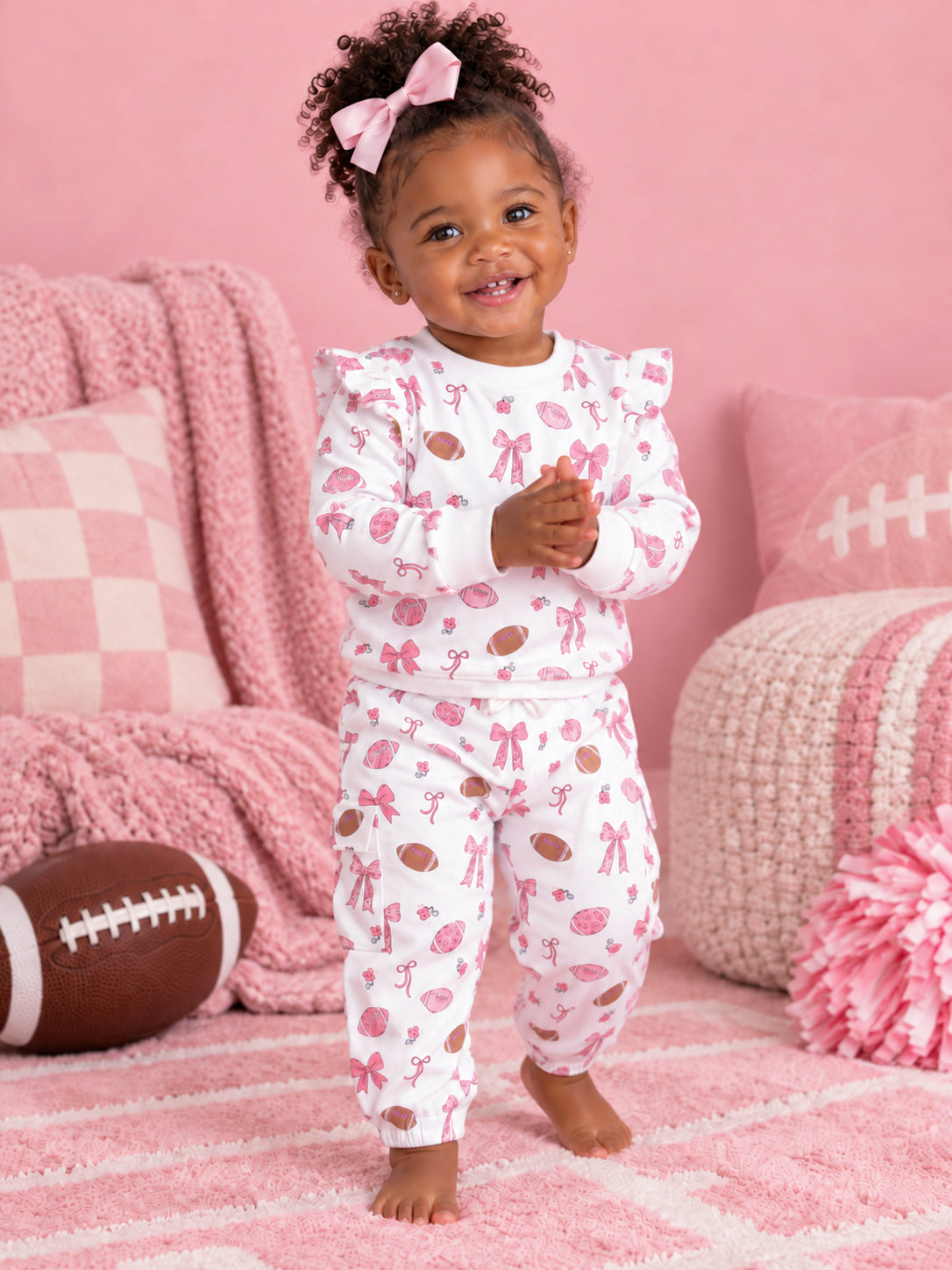 Toddler in pink patterned pajamas smiles, playing indoors with a football, surrounded by soft pink decor.