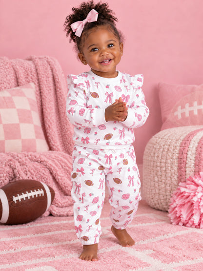 Toddler in pink patterned pajamas smiles, playing indoors with a football, surrounded by soft pink decor.