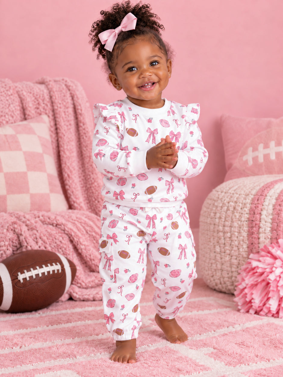 Toddler in pink patterned pajamas smiles, playing indoors with a football, surrounded by soft pink decor.