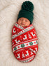 Newborn swaddled in festive red pattern with reindeer, wearing a green pom-pom hat, resting on a plush white blanket.
