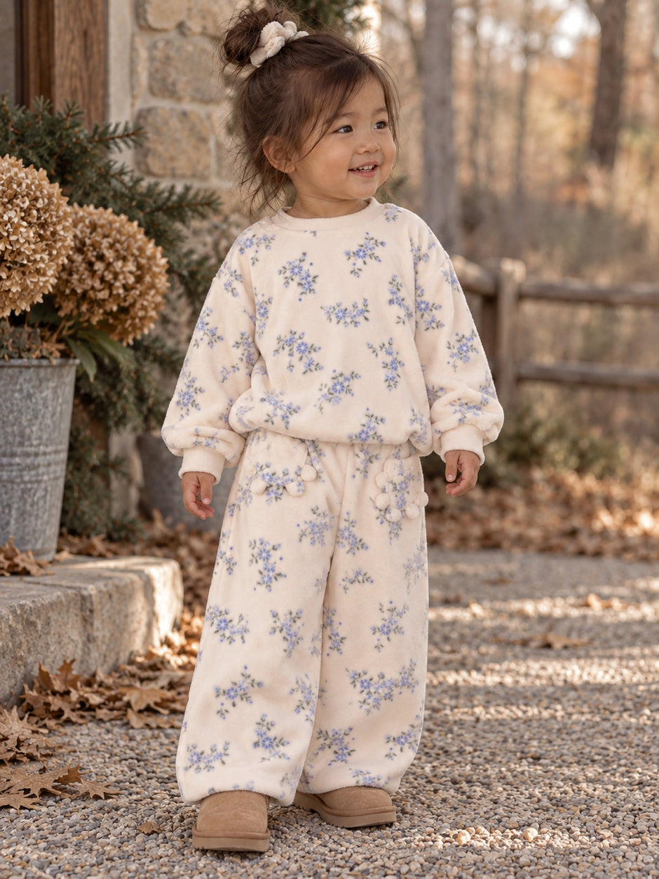 Child in floral loungewear smiles outdoors, surrounded by plants and autumn leaves. Cozy and comfortable outfit.