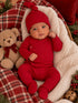 Infant in a red outfit and hat sits with a teddy bear on a cozy blanket amidst festive decor.