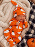 Smiling baby in an orange ghost-themed onesie and hat, resting peacefully on a cozy blanket with a pumpkin nearby.