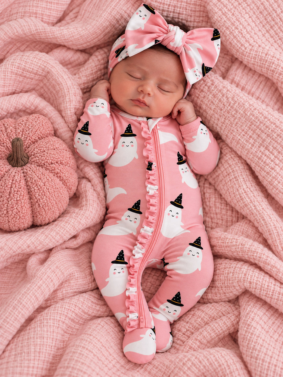 Baby girl in pink ghost-patterned onesie and matching headband, resting on a pink blanket with a pumpkin.