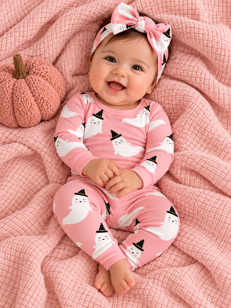 Smiling baby in pink ghost-themed pajamas sits on a soft blanket with a pumpkin, wearing a matching headband.