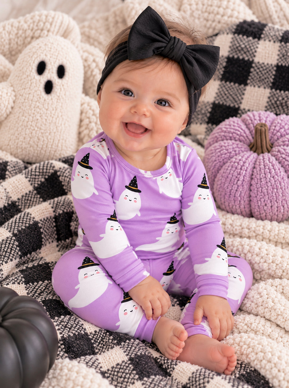 Smiling baby in purple ghost pajamas with a black bow, surrounded by Halloween decorations and cozy blankets.