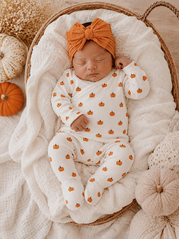 Infant sleeping peacefully in a cozy basket, wearing a white pumpkin-patterned outfit and a large orange headband.