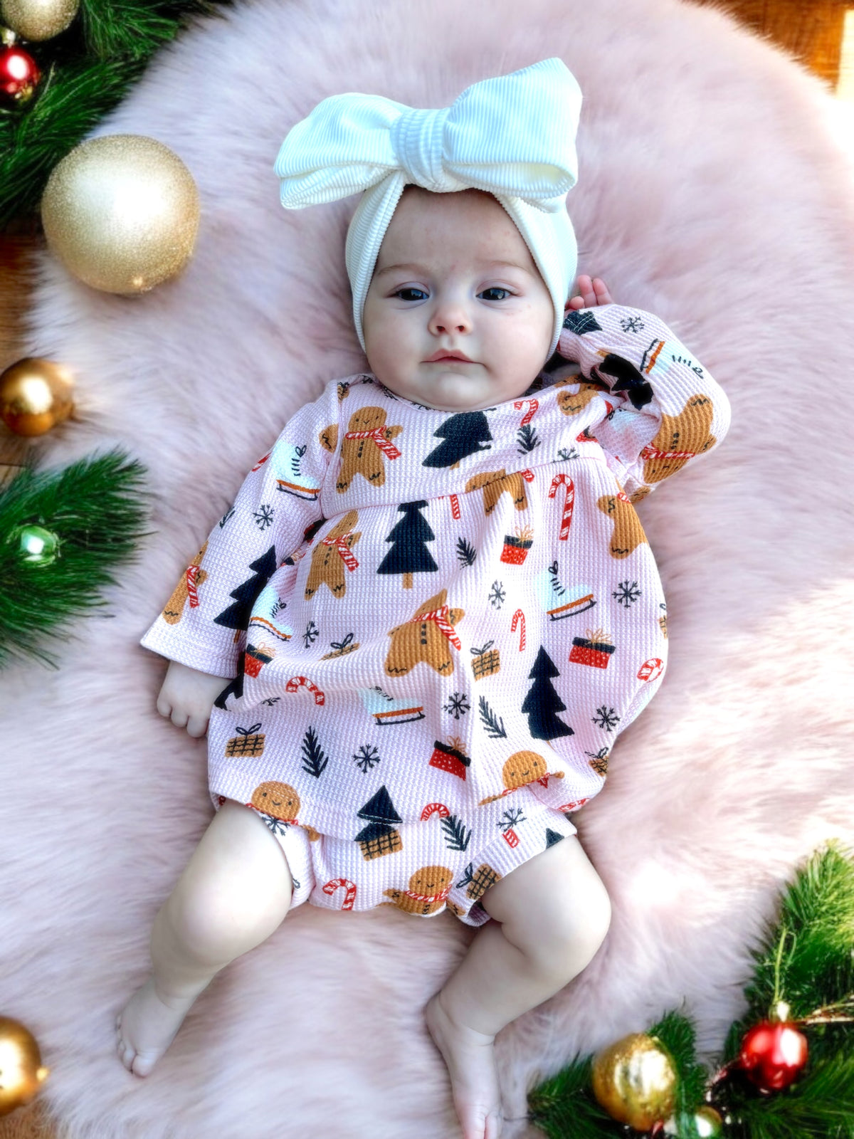Baby in festive outfit with gingerbread and Christmas tree patterns, lying on a pink plush background with holiday decor.