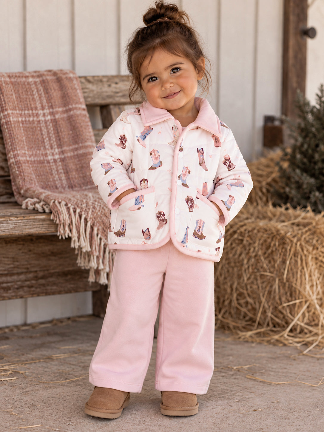 Smiling toddler in a patterned jacket and pink pants, sitting on a rustic bench with a cozy blanket nearby.