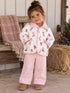 Smiling toddler in a patterned jacket and pink pants, sitting on a rustic bench with a cozy blanket nearby.