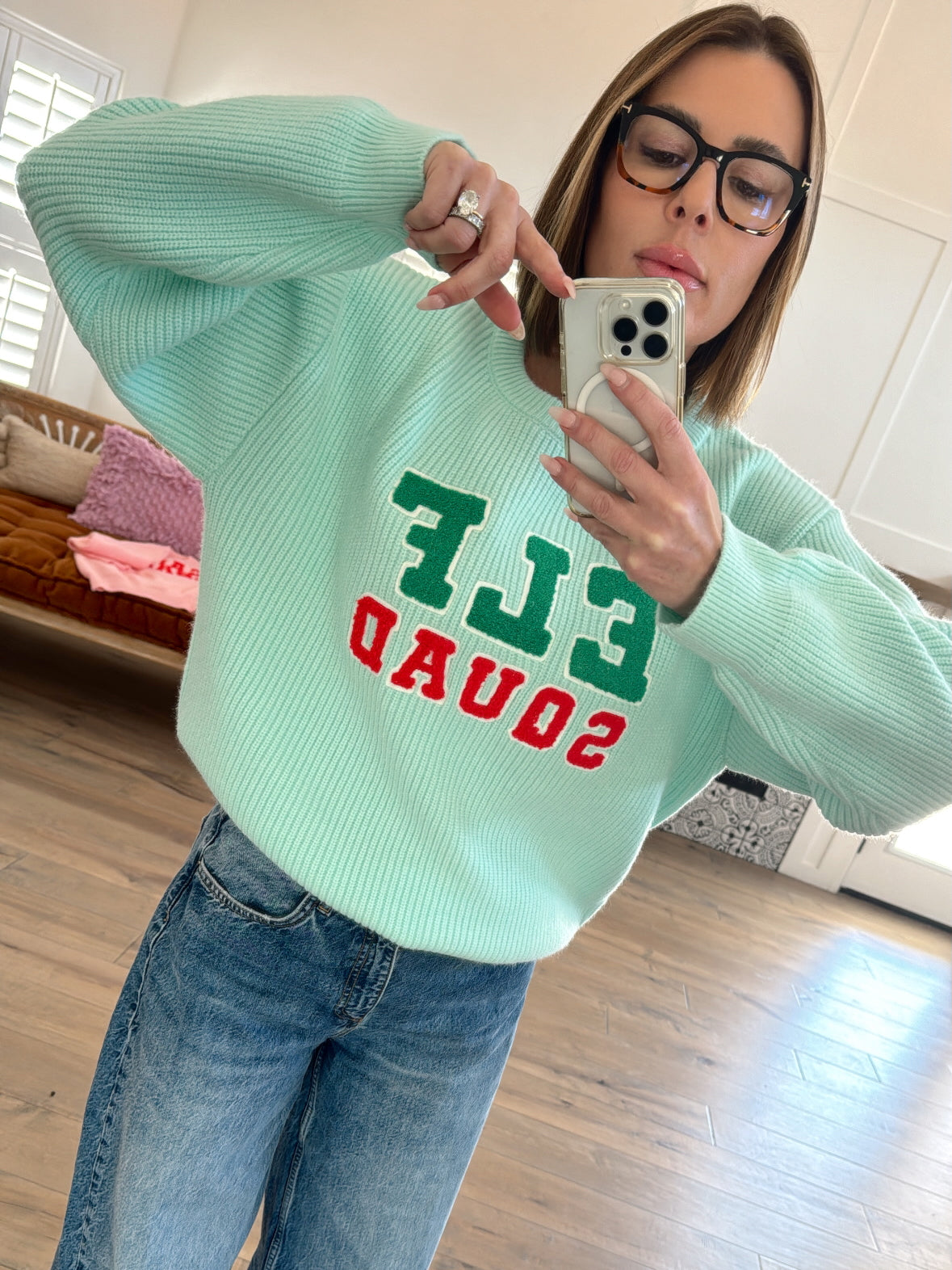 Person wearing a mint green sweater with "ELF SQUAD" text, posing while taking a selfie in a cozy indoor setting.