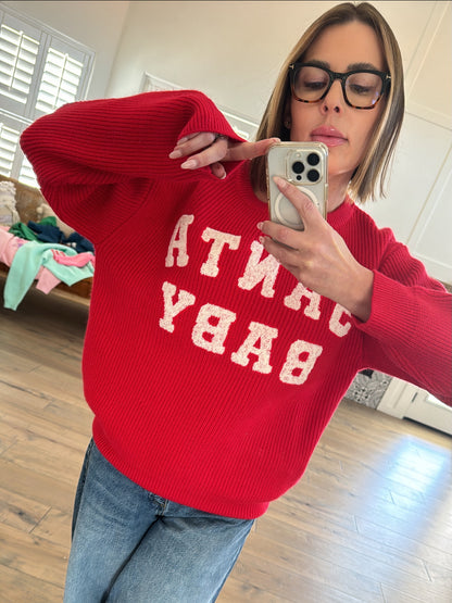 Person wearing a red sweater with "SANTA BABY" printed, posing and taking a selfie in a bright room.