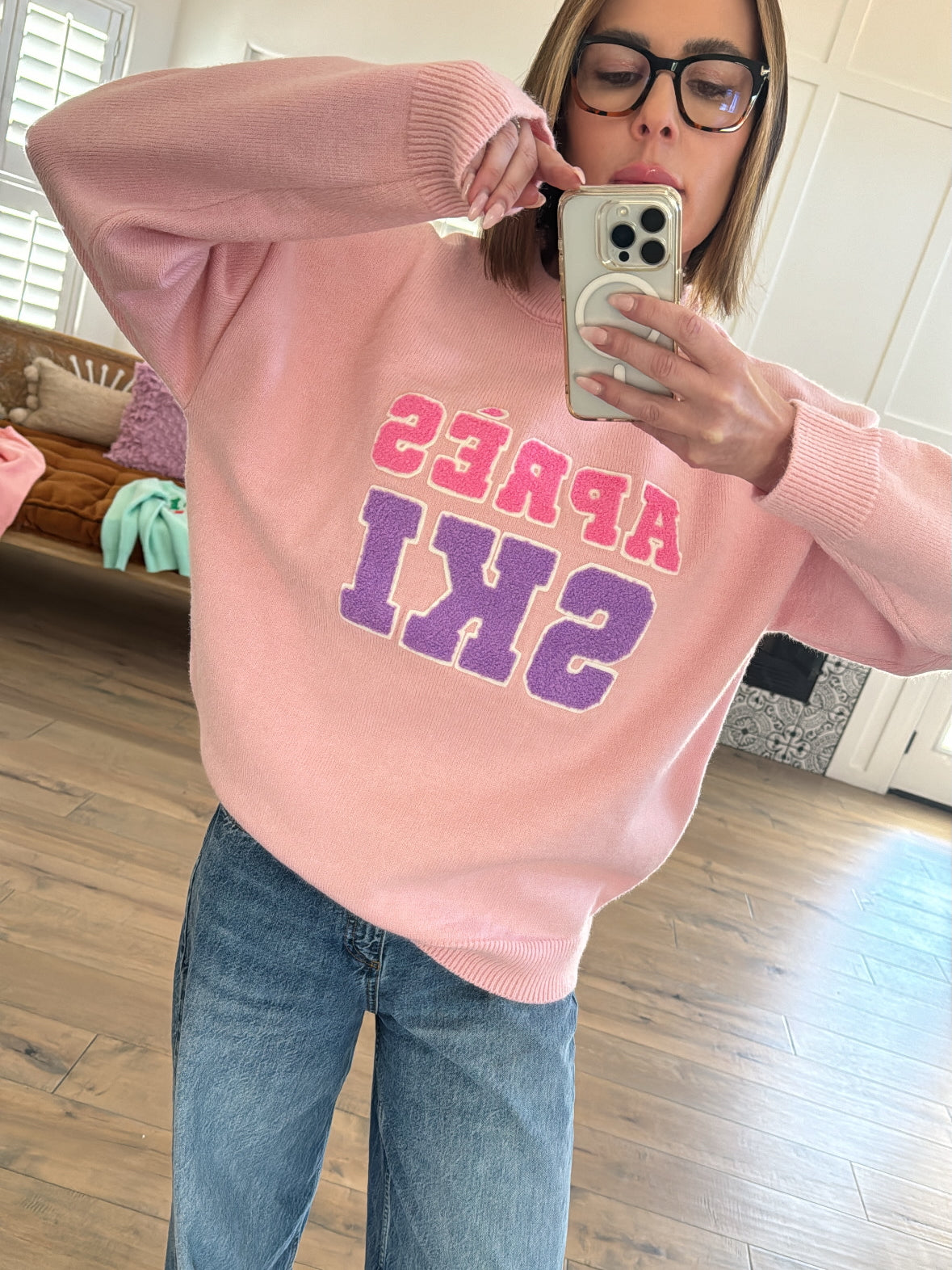 Woman wearing a pink sweater with "APRES SKI" text, taking a selfie in a bright, cozy room.