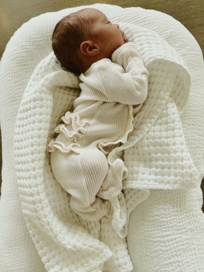 Newborn sleeping peacefully on a soft, textured white blanket, wrapped snugly in a light-colored outfit.