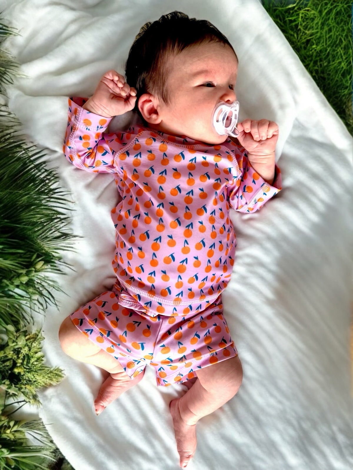 Baby lying on a white blanket, wearing a colorful outfit with fruit patterns and a pacifier in their mouth.