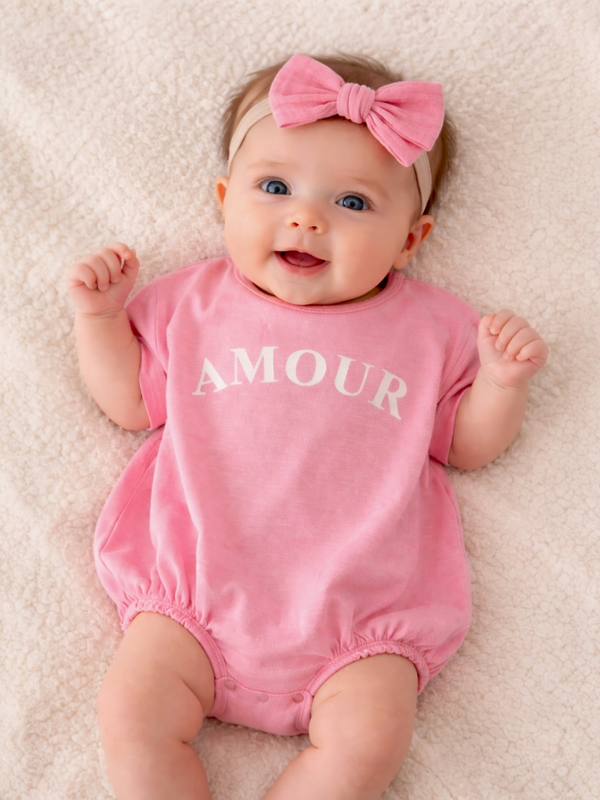 Smiling baby in a pink onesie with "AMOUR" printed, wearing a matching pink bow, on a soft blanket.