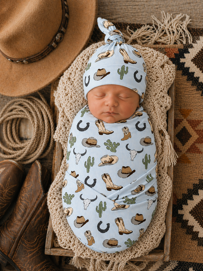 Newborn swaddled in a blue western-themed wrap, resting on a textured blanket with cowboy boots and hat nearby.
