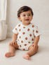 Smiling baby in a floral-patterned outfit, seated on a soft surface with natural light.