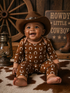 Smiling baby in a cowboy hat, wearing a brown outfit with stars and horseshoes, sitting on a patterned rug.