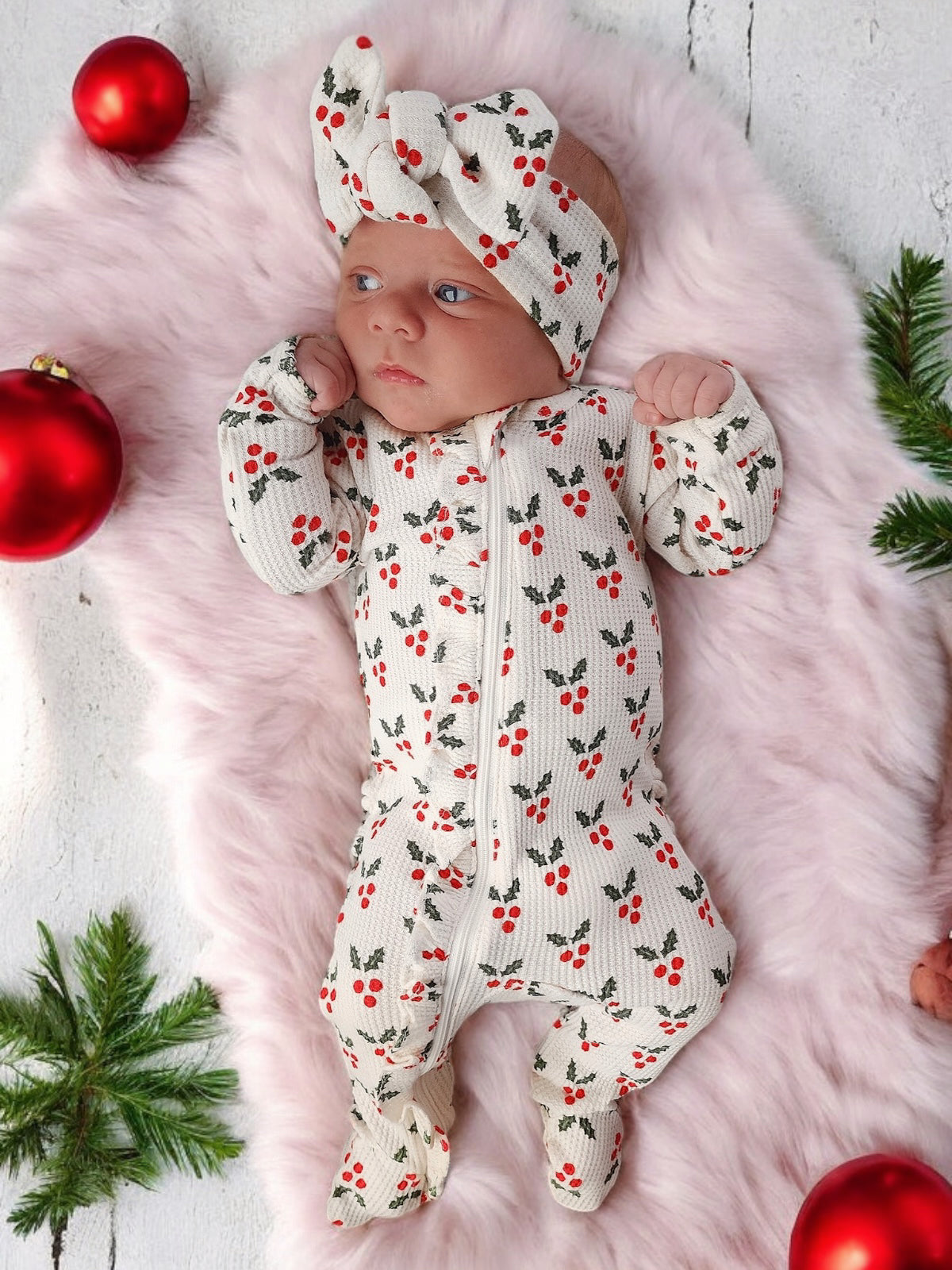Baby in holiday-themed outfit with cherry pattern, wearing a matching headband, on fluffy blanket surrounded by ornaments.