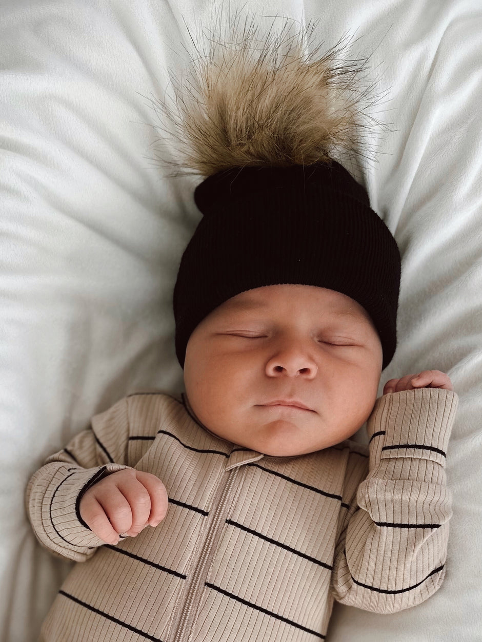 Sleeping baby in a cozy striped onesie and a black beanie with a fluffy pom-pom, lying on a white blanket.