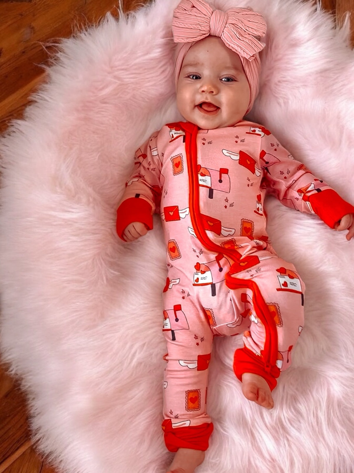 Happy baby in pink pajamas with hearts, smiling on a fluffy blanket with a matching headband.