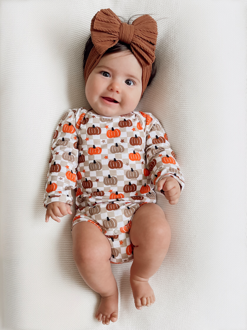 Smiling baby on a textured blanket, wearing a pumpkin-patterned outfit and a large brown headband.