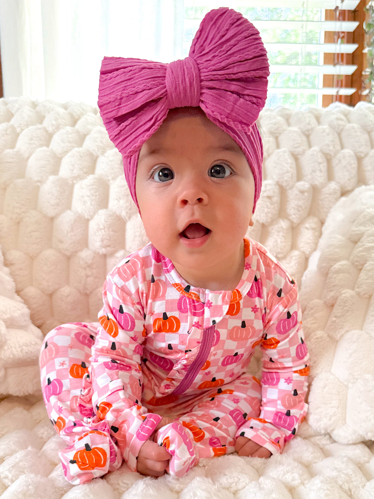 Baby in pink pumpkin onesie with a large pink bow, sitting on a soft, textured blanket. Bright, cheerful expression.