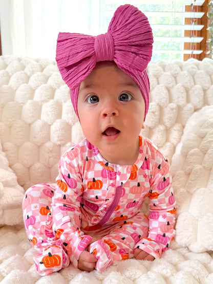Baby in pink pumpkin onesie with a large pink bow, sitting on a soft, textured blanket. Bright, cheerful expression.
