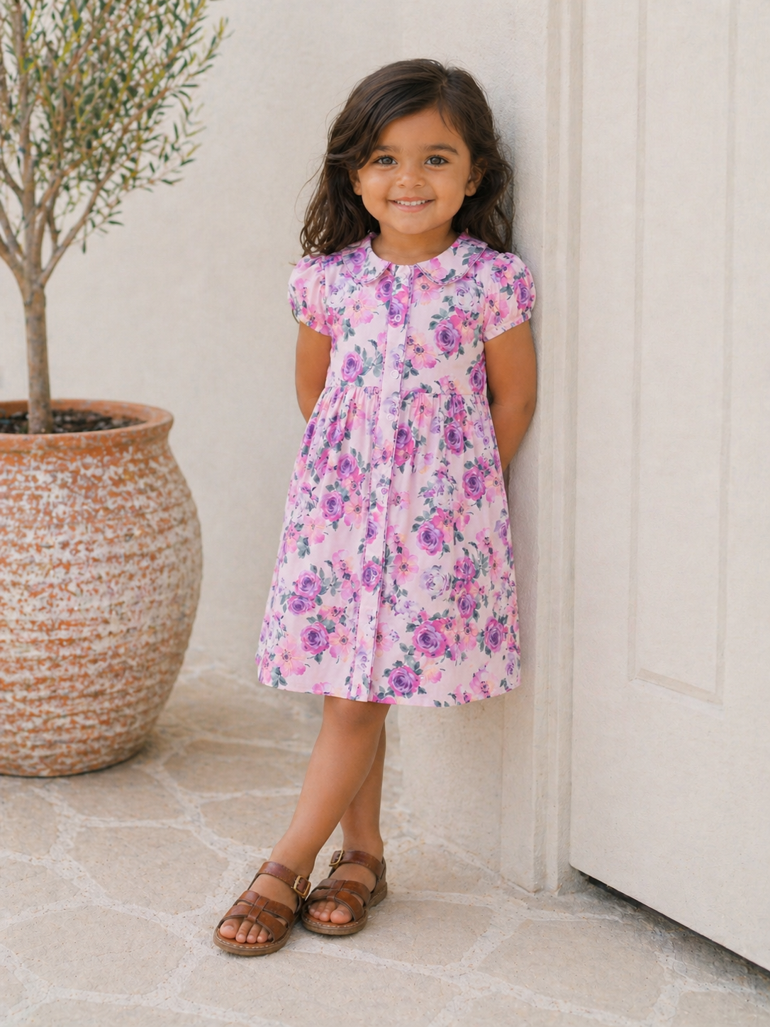 Smiling girl in a floral pink dress stands beside a pot plant against a light wall. Sunny, cheerful ambiance.